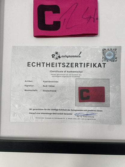 Brazalete de capitán firmado por Rudi Völler en un marco de autógrafos con temática alemana.