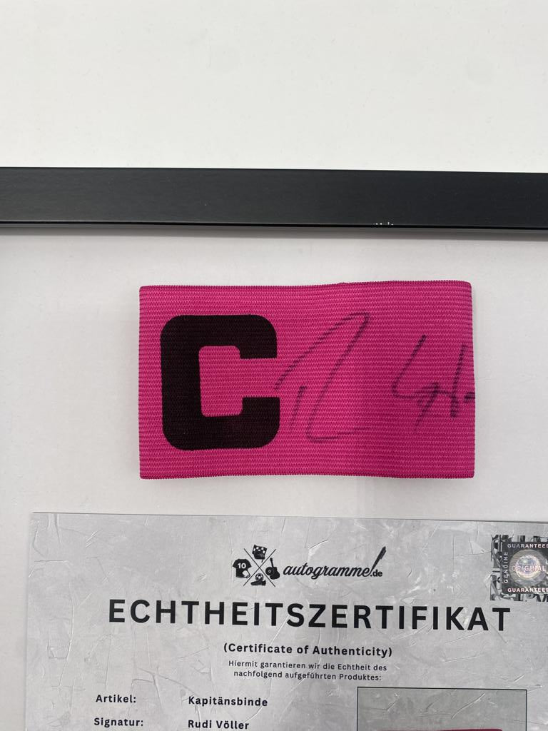 Brazalete de capitán firmado por Rudi Völler en un marco de autógrafos con temática alemana.