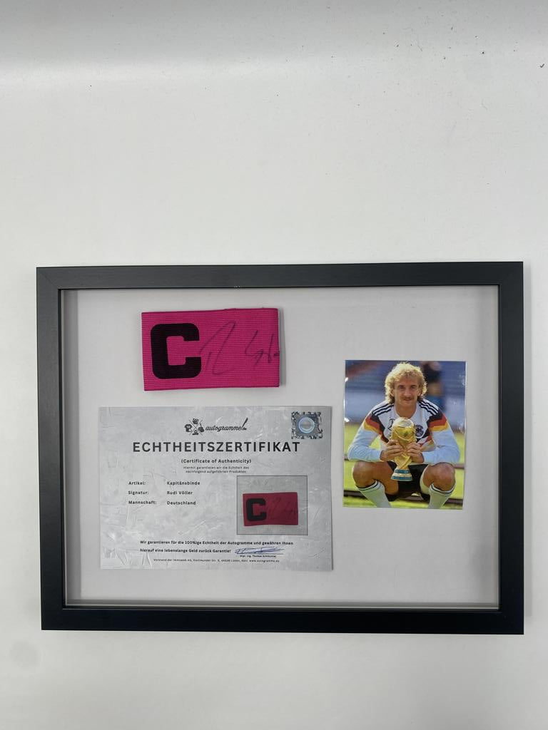Brazalete de capitán firmado por Rudi Völler en un marco de autógrafos con temática alemana.